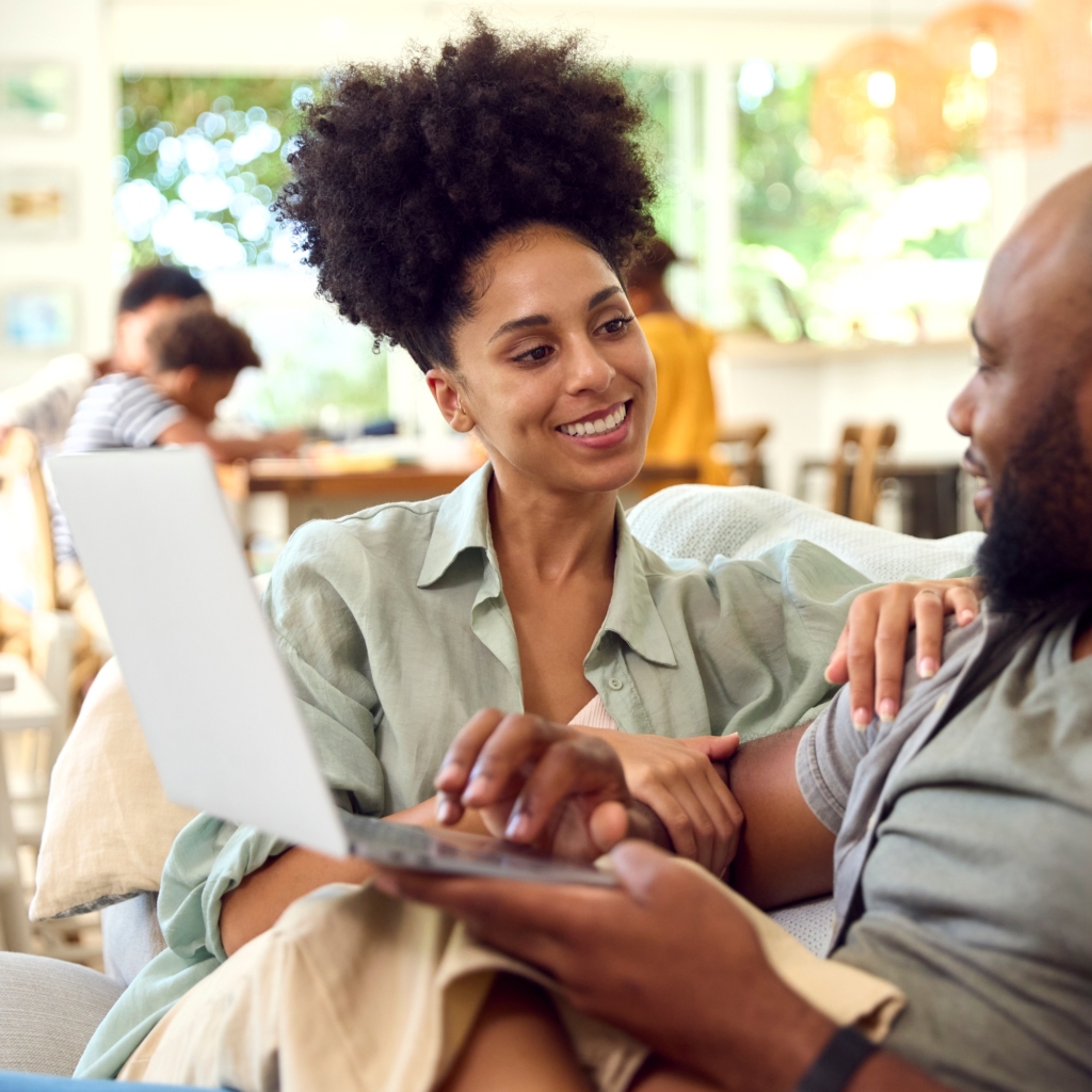 Couple At Home Using Laptop To Shop Or Book Holiday With Multi-Generation Family In Background