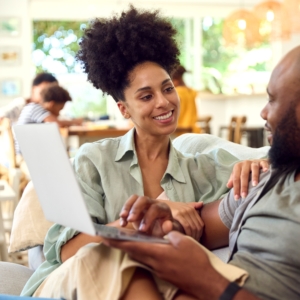 Couple At Home Using Laptop To Shop Or Book Holiday With Multi-Generation Family In Background